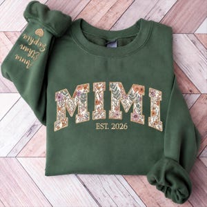 Könnte beinhalten: Ein olivgrüner Rundhals-Pullover mit dem Wort MIMI in großen Blockbuchstaben, die mit einem bunten Blumenmuster gefüllt sind. Unter dem Text steht der Schriftzug EST. 2026. Der linke Ärmel ist mit den Namen Anna, Ethan und Sophia bestickt.