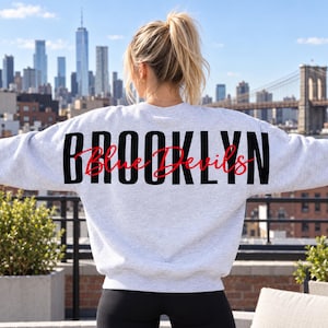 Könnte beinhalten: Ein hellgraues Sweatshirt mit Rundhalsausschnitt, das auf der Rückseite das Wort BROOKLYN in großen schwarzen Großbuchstaben zeigt, mit dem in roter Schreibschrift geschriebenen Text Blue Devils über dem schwarzen Text.
