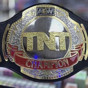 May include: A black leather championship wrestling belt featuring a large central metal plate with gold-colored TNT lettering, a red banner with the word CHAMPION, and an AEW logo at the top. The plate has silver textured accents and side panels.