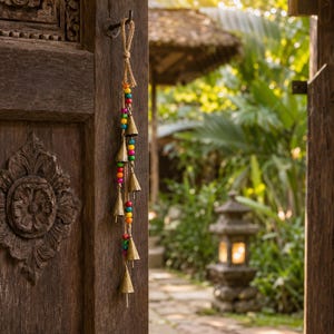 May include: A hanging wind chime featuring five conical brass bells suspended by a jute twine cord. The chime is decorated with a vertical arrangement of small, round, multi-coloured beads in shades of red, orange, yellow, green, and blue.