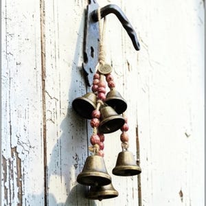 Può includere: Un ornamento sospeso con cinque campanelle in metallo color ottone appese a uno spago di juta. La composizione include piccole perline di legno rotonde color marrone rossiccio screziato e un ciondolo metallico circolare, fissato a un gancio.
