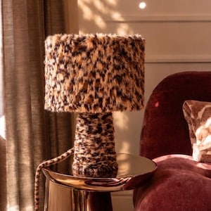 May include: A table lamp featuring a cylindrical base and a drum shade, both covered in a brown and black leopard print faux fur fabric. The lamp sits on a polished, reflective copper-toned metal side table with a smooth, tapered cylindrical base.