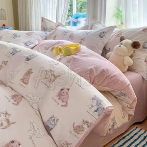 May include: A bedding set featuring a light pink and white colour scheme with a repeating pattern of illustrated kittens in various poses. The set includes a duvet cover and pillowcases, accented by a plush dog toy and yellow headphones.