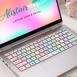 Könnte beinhalten: Ein silberner Laptop mit einer Tastaturabdeckung in Pastellrosa, Blau und Gelb, verziert mit kleinen Zeichentrickfiguren. Der Bildschirm zeigt einen Farbverlauf mit dem Text AlistairShopArt LAPTOP SKIN & DECAL.