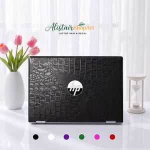 Op de afbeelding: Een zwarte laptopsticker met een textuur van krokodillenleer op de klep van een HP-laptop. In het midden staat het zilveren HP-logo. Onder de laptop staan zes kleine gekleurde cirkels in zwart, wit, paars, groen, roze en rood.