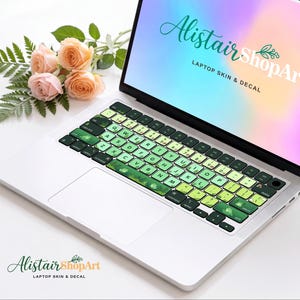 Peut inclure: Un autocollant pour clavier d'ordinateur portable avec un dégradé de touches vert clair à vert foncé, décoré de petites illustrations de grenouilles et de plantes. L'écran affiche le texte AlistairShopArt LAPTOP SKIN & DECAL sur un fond coloré.