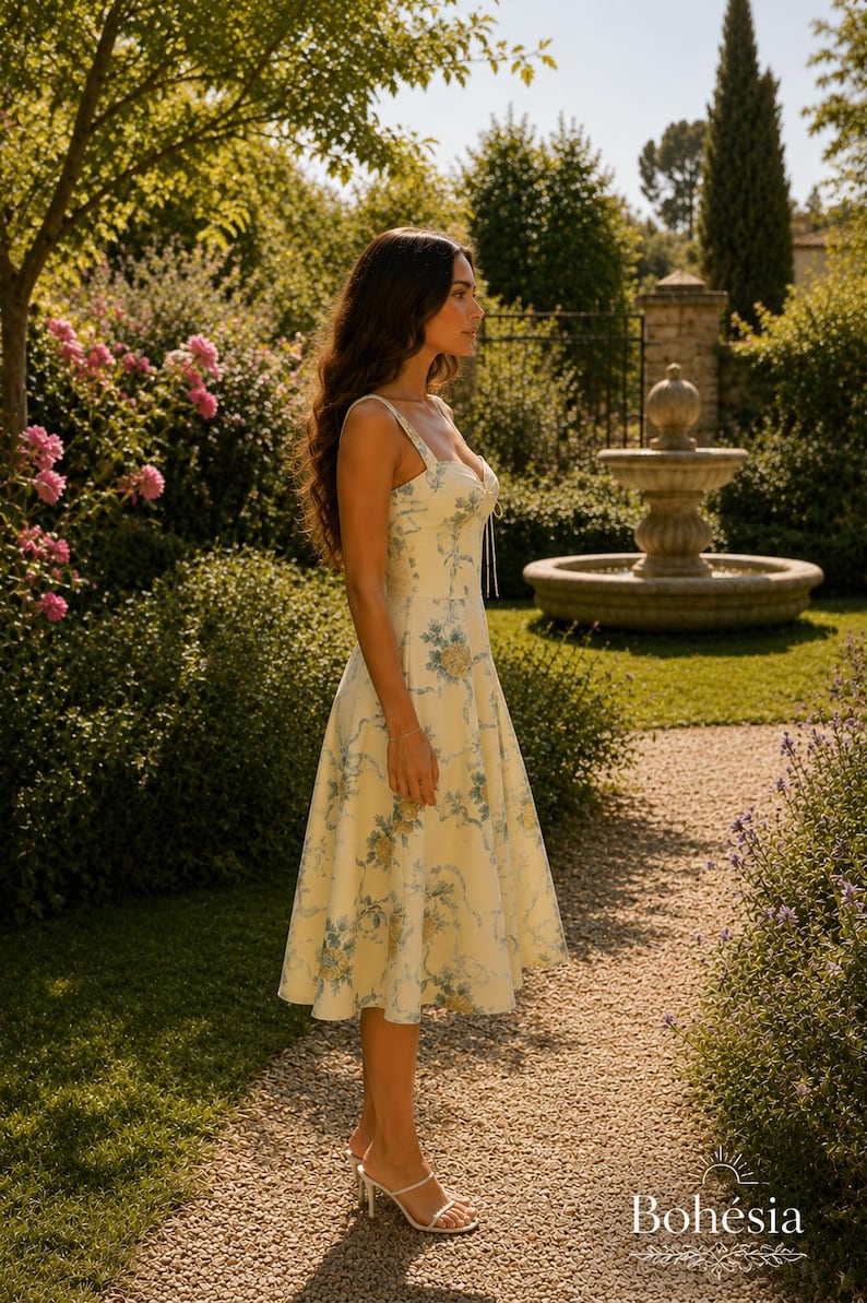 May include: A cream-colored midi dress featuring a blue and yellow floral pattern, a sweetheart neckline, and thin shoulder straps. The dress has a fitted bodice and a flared skirt that falls below the knees, paired with white strappy sandals.