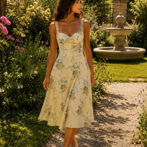 May include: A midi-length dress in a pale yellow hue featuring a blue and yellow floral pattern with ribbon motifs. The garment has a sweetheart neckline, thick shoulder straps, a fitted bodice with a central tie detail, and a flared skirt.