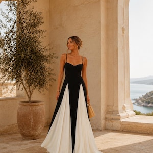 Peut inclure: Une robe longue au sol avec un corsage noir à fines bretelles et un décolleté en cœur, contrastée par des panneaux plissés blancs fluides intégrés à la jupe. Le mot BOHÉSIA est imprimé dans le coin inférieur droit de l'image.