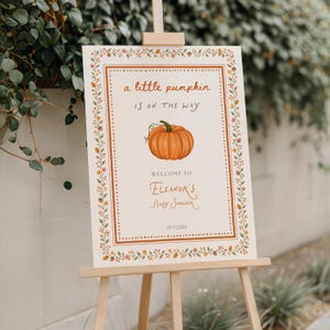 Puede incluir: Un letrero rectangular sobre un caballete de madera con el texto 'a little pumpkin IS ON THE WAY', una ilustración central de una calabaza naranja y el texto 'WELCOME TO Eleanor's Baby Shower' con la fecha '07.11.2026' dentro de un borde otoñal.