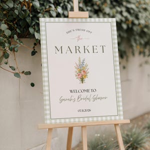 Puede incluir: Un letrero rectangular con un borde de cuadros vichy en verde claro y blanco sobre un caballete de madera. El letrero presenta el texto "SHE'S FRESH OFF THE MARKET", "WELCOME TO Sarah's Bridal Shower" y la fecha "07.11.2026" con una ilustración floral central.