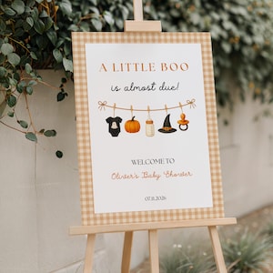 Puede incluir: Un letrero rectangular para baby shower sobre un caballete de madera con borde de cuadros beige. El centro blanco presenta el texto A LITTLE BOO is almost due!, una ilustración de artículos de bebé en un tendedero y 07.11.2026.