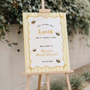 Puede incluir: Un letrero rectangular con un borde de rayas amarillas y blancas que presenta ilustraciones de abejas y el texto: She found her honey and it's meant to bee! Welcome to Eleanor's Bridal Shower! 09.14.2026. El letrero se muestra en un caballete de madera.