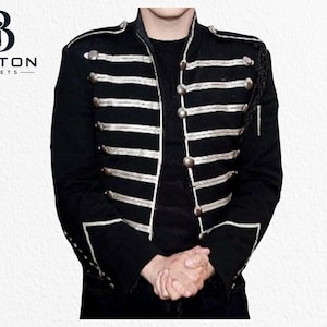 Könnte beinhalten: Eine schwarze Jacke im Militärstil mit silberfarbenen horizontalen Zierborten auf der Brust und silbernen Knöpfen. Die Jacke hat einen Stehkragen, strukturierte Schultern mit Schulterklappen sowie weiße Paspeln an den Ärmelbündchen und der Vorderseite.