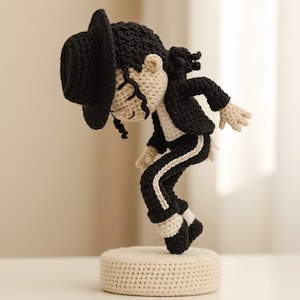 Könnte beinhalten: Eine gehäkelte Figur eines Künstlers mit schwarzem Fedora, schwarzer Jacke und schwarzer Hose mit weißem Seitenstreifen. Die Figur hat beige Haut, lockiges schwarzes Haar und steht in einer nach vorne geneigten Pose auf einem runden, beigen Sockel.