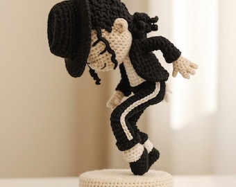 May include: A crocheted figurine of a performer in a black fedora, black jacket, and black trousers with a white side stripe. The figure has beige skin, curly black hair, and is posed in a forward-leaning stance on a round, beige crocheted base.