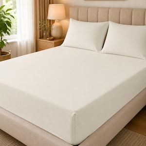 May include: A bed featuring a smooth, solid off-white fitted sheet covering the mattress, paired with two matching off-white rectangular pillows placed against a tufted, light beige upholstered headboard in a neutral bedroom setting.
