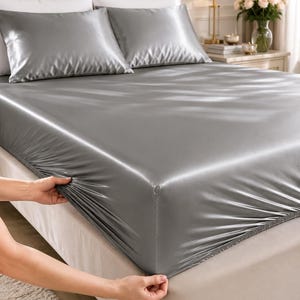 May include: A set of solid grey satin bed sheets featuring a fitted sheet with elasticized corners and two matching pillowcases. The fabric has a smooth, reflective finish and a lustrous sheen, covering a mattress on a tufted headboard bed frame.