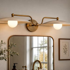 Puede incluir: Un aplique de pared de dos luces con estructura de metal en acabado latón y una placa de montaje circular central. Dos pantallas anchas y poco profundas en forma de platillo de color blanco roto se extienden horizontalmente, cada una con una bombilla esférica de cristal blanco debajo.