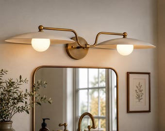Può includere: Una lampada da parete a due luci con struttura in ottone e due paralumi larghi e poco profondi color bianco sporco. Ogni paralume ospita una lampadina sferica in vetro smerigliato che emette una luce calda, montata su una base circolare.