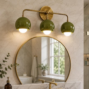 Può includere: Una lampada da parete a tre luci con struttura in ottone spazzolato e tre paralumi sferici in vetro verde oliva. Ogni paralume contiene una lampadina bianca luminosa, montata su una piastra circolare in ottone.