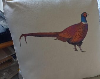 May include: Cream-colored decorative pillow featuring a detailed illustration of a pheasant. The pheasant has a blue head, orange and brown body, and a long tail. The pillow has a simple, clean design.