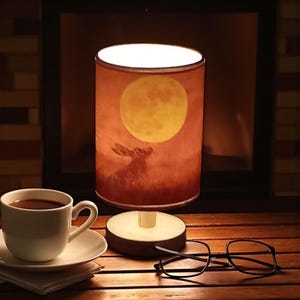 Puede incluir: Lámpara de mesa cilíndrica con una pantalla de color marrón rojizo que representa una luna llena y una silueta de conejo. La lámpara está sobre una base de madera. Una taza de café, un platillo y gafas están sobre una mesa de madera.