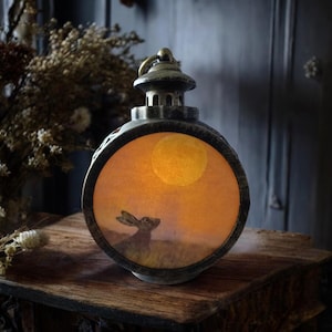 May include: A bronze-finished decorative lantern with a circular window. Inside, a rabbit gazes at a full moon in an orange sky. The lantern is placed on a wooden surface. The overall theme is a rustic, nighttime scene.