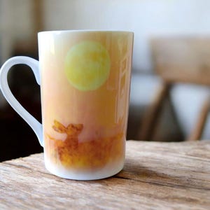 Può includere: Una tazza in ceramica bianca con una scena di tramonto con una luna gialla e una silhouette di cervo. La tazza ha un manico arrotondato.
