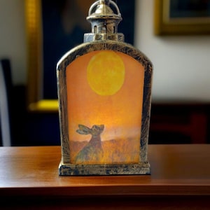 Könnte beinhalten: Eine dekorative Laterne mit goldfarbenem Rahmen und einem Griff oben. Die Laterne zeigt ein Gemälde eines Hasen, der einen großen gelben Mond in einem orangefarbenen Himmel betrachtet. Die Laterne steht auf einer Holzoberfläche.