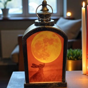 Peut inclure: Une lanterne décorative avec un cadre en métal et une poignée. La lanterne présente une image rétro-éclairée d'un lapin regardant une pleine lune. La palette de couleurs est chaude, avec des tons orange et marron. Des bougies sont allumées en arrière-plan.