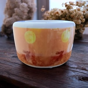 Puede incluir: Una taza de cerámica blanca con una escena de puesta de sol y dos siluetas de ciervos. La taza tiene un fondo naranja claro con dos círculos amarillos que representan el sol.