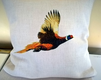 May include: Square decorative pillow with a detailed image of a pheasant in flight. The bird features brown, orange, and yellow feathers, with a red and blue head. The pillow has a neutral, off-white background.