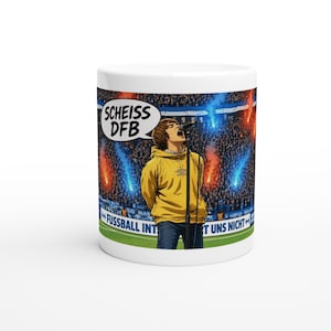Op de afbeelding: Een witte keramische mok met een grafische illustratie van een persoon in een gele hoodie die in een microfoon zingt. De achtergrond toont een vol stadion met blauwe en rode fakkels. Tekst: SCHEISS DFB en FUSSBALL INTERESSIERT UNS NICHT.