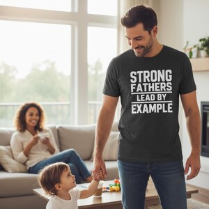 May include: A dark charcoal gray short-sleeved t-shirt featuring the white text "STRONG FATHERS LEAD BY EXAMPLE" printed in a bold, block-style font across the chest, with horizontal lines separating the words for a clear, graphic design.