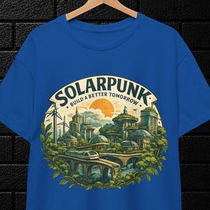 May include: A royal blue t-shirt featuring a circular graphic print of a futuristic city with lush greenery, wind turbines, and a train on a bridge. The text reads "SOLARPUNK" and "BUILD A BETTER TOMORROW" in a curved font above the illustration.
