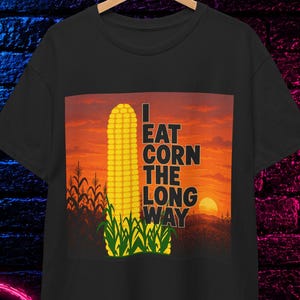 Può includere: Una t-shirt nera a maniche corte con una stampa grafica di una pannocchia di mais gialla su uno sfondo al tramonto. Il testo "I EAT CORN THE LONG WAY" è stampato in lettere maiuscole nere accanto al mais. La maglietta è appesa a una gruccia di legno.