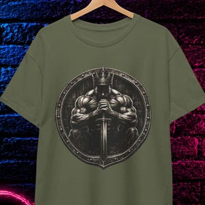 May include: An olive green t-shirt featuring a circular graphic of a muscular figure wearing a crown and holding a sword. The shirt is displayed on a wooden hanger and includes a circular badge in the corner that reads 100% COTTON.