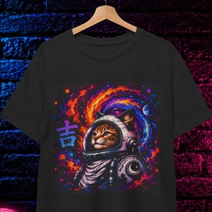 May include: A black t-shirt featuring a graphic print of a cat wearing a space helmet against a colorful swirling galaxy background. The design includes the purple Japanese character 吉 on the left side. The shirt is displayed on a wooden hanger.