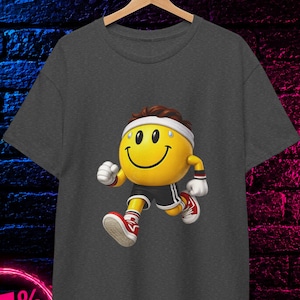 Op de afbeelding: Een antracietgrijs t-shirt met een afbeelding van een geel smiley-personage met een witte hoofdband, zwarte sportbroek en rode sneakers terwijl het rent. Het shirt bevat een cirkelvormige badge in de hoek met de tekst 100% COTTON.