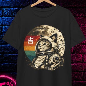 Op de afbeelding: Een zwart t-shirt met een afbeelding van een kat in een astronautenhelm tegen een maanachtergrond. Het ontwerp bevat een retro-cirkel met horizontale strepen in rood, oranje en blauwgroen, samen met het Japanse karakter 吉 in wit.