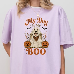 Peut inclure: Un t-shirt à manches courtes couleur orchidée avec un motif représentant un chien déguisé en fantôme portant un bandana à carreaux. Le design inclut deux citrouilles d'Halloween, des chauves-souris et le texte "My Dog is My BOO" en brun et orange.