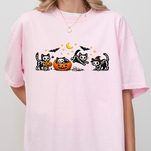 May include: A light pink short-sleeve t-shirt featuring a graphic print of four black skeleton cats, one sitting inside an orange pumpkin, surrounded by small yellow stars, a crescent moon, and black bats. The shirt has a crew neckline.