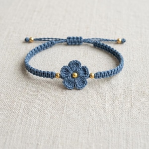 May include: A blue macrame bracelet featuring a central crocheted flower motif. The design includes a small gold-toned bead at the center of the flower and two additional gold-toned beads positioned on the cord on either side of the flower.