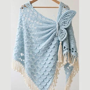May include: A light blue crochet shawl featuring a prominent butterfly motif on one side. The garment is constructed with a scalloped stitch pattern and is finished with cream-colored fringe and small white beads along the bottom edge.