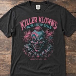 以下が含まれることがあります： フリル付きの襟のピエロのグラフィックが描かれた黒の半袖Tシャツ。ピンクと青で「KILLER KLOWNS FROM OUTER SPACE」と書かれており、下部には小さなピンクの文字で「CRESCENT COVE, CA - 1988」とプリントされています。
