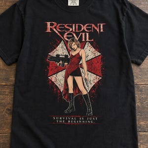 May include: A black short-sleeved t-shirt featuring a graphic of a character in a red dress holding a firearm, set against a red and white umbrella logo. The text RESIDENT EVIL and SURVIVAL IS JUST THE BEGINNING are printed in white and red.