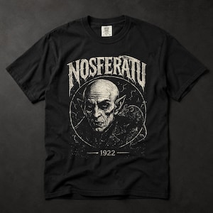 Puede incluir: Una camiseta negra de manga corta con un estampado gráfico blanco desgastado del personaje Nosferatu de la película de 1922. El diseño incluye el texto NOSFERATU arqueado sobre el retrato y el año 1922 centrado debajo del marco circular.