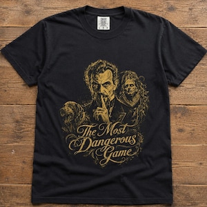 Könnte beinhalten: Ein schwarzes Kurzarm-T-Shirt mit einem goldenen Grafikdruck eines Mannes, der einen Finger auf die Lippen legt, sowie einem Hund und einer schattenhaften Figur. Der Text "The Most Dangerous Game" steht in dekorativer Schrift unter dem Motiv.