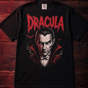 Puede incluir: Una camiseta negra de manga corta con un estampado gráfico de Drácula en rojo, blanco y negro. La palabra DRACULA está impresa en una fuente roja irregular sobre el retrato, que muestra una figura con esmoquin de cuello alto y murciélagos.